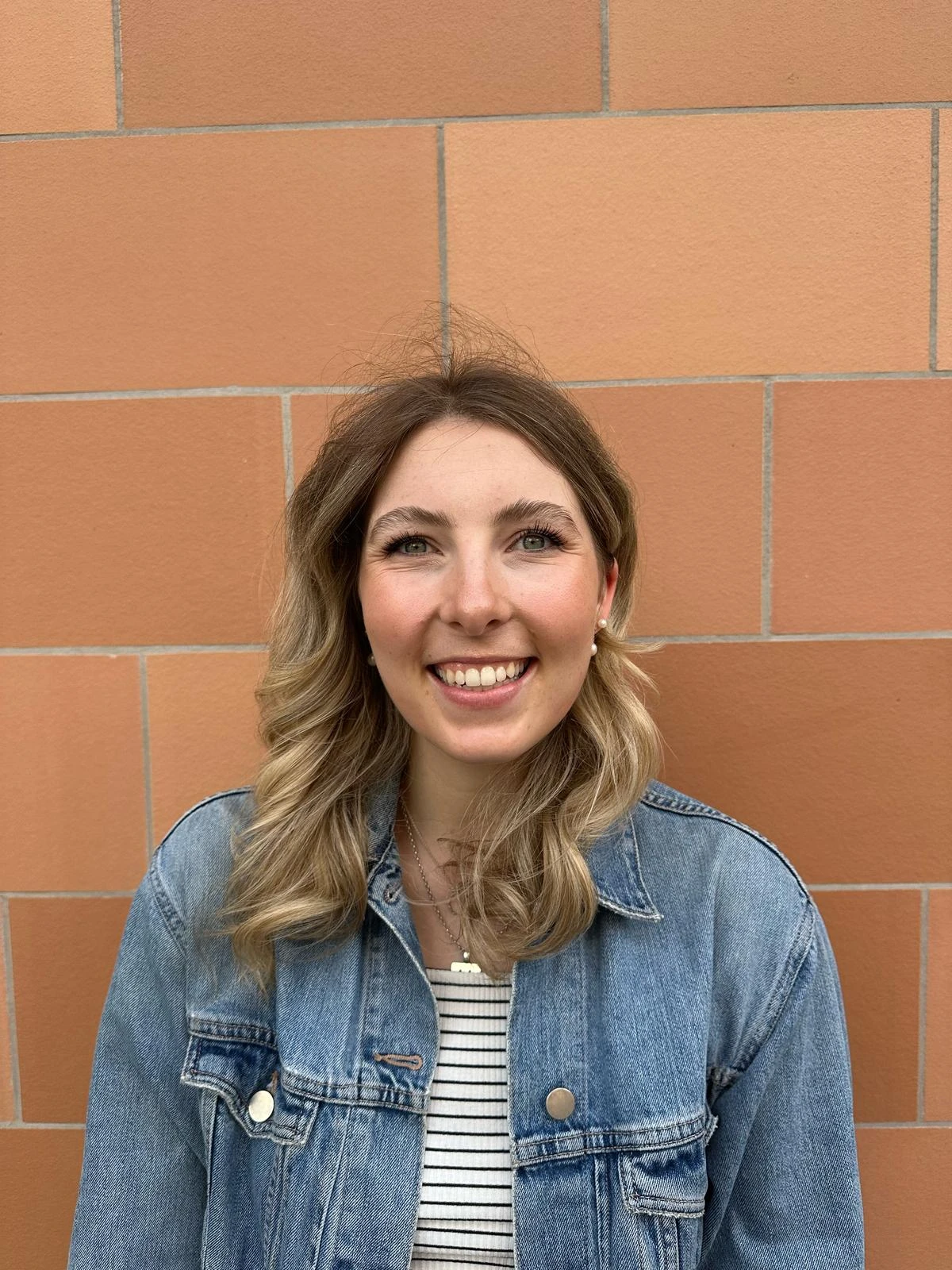 Smiling woman with shoulder-length hair wearing a denim jacket against a brick wall.