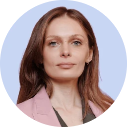 A woman with long, wavy brown hair and fair skin, wearing a pink blazer and a neutral expression.
