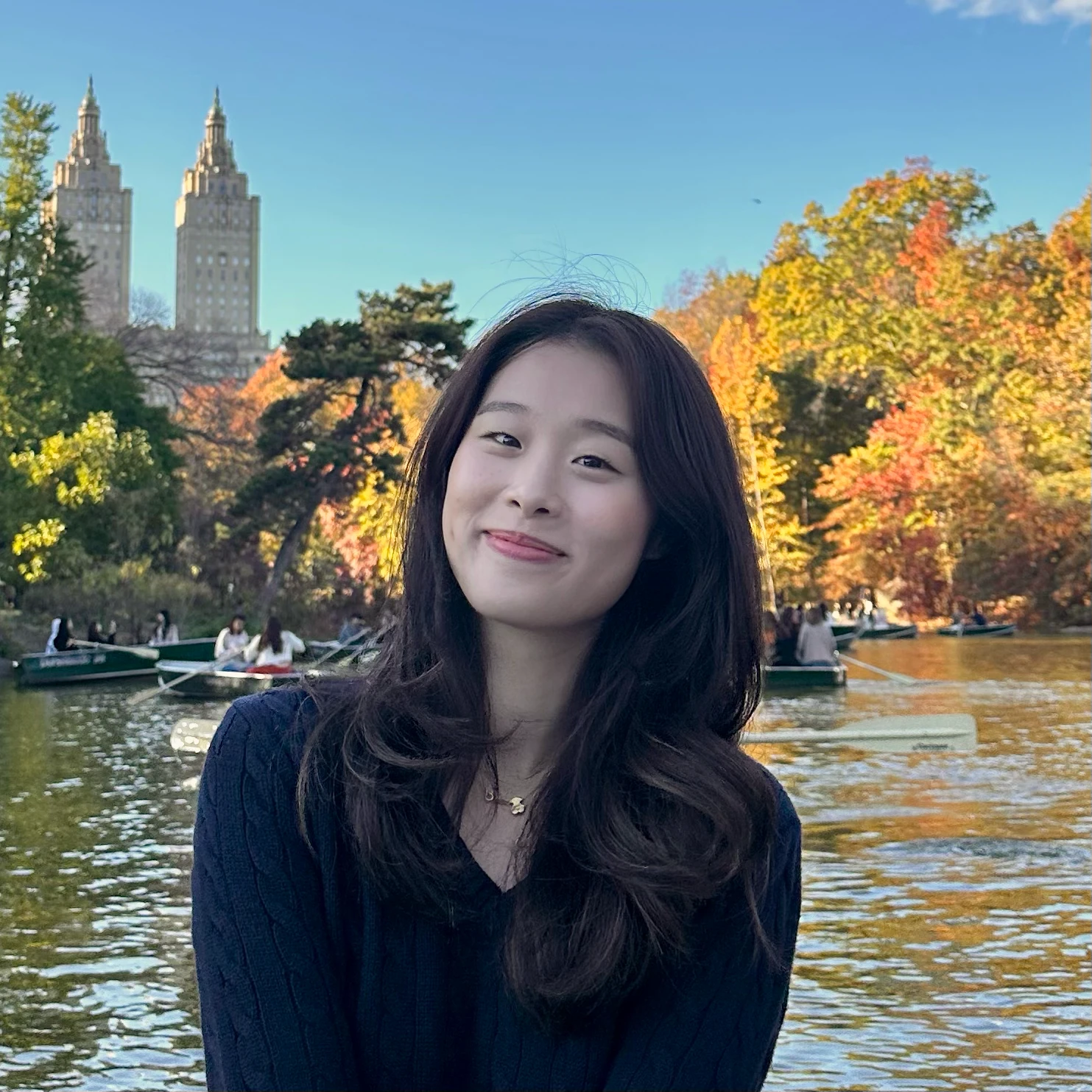 A smiling woman with long hair at a lake surrounded by autumn trees and boats.